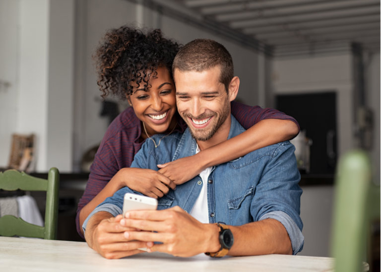 An interracial couple at a desk, looking at a mobile phone together and smiling 